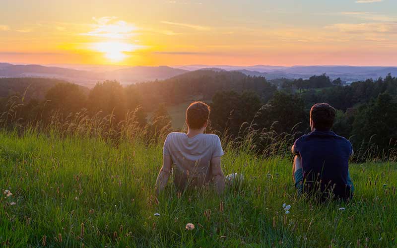 Two men sit shoulder-to-shoulder and watch the sun set over a field.